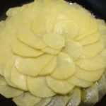 Patate finemente affettate, adagiate in padella come secondo strato nella preparazione della ricetta interamente vegetale di patate e cipolle in padella