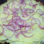 Patate finemente affettate, adagiate in padella con cipolle finemente tagliate per la preparazione della ricetta interamente vegetale di patate e cipolle in padella